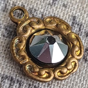 May include: A gold-toned metal pendant with a decorative, ornate design. The pendant features a large, faceted, clear glass stone in the center.