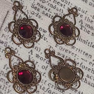 May include: Four antique-style brass pendants with a floral design. Each pendant features a dark red, oval-shaped gemstone in the center, surrounded by an ornate, openwork frame. One pendant has an empty setting. The pendants are for jewelry making.