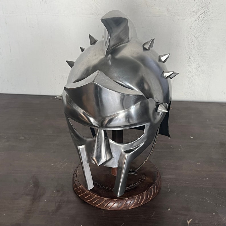 May include: A polished silver gladiator helmet with a prominent brow guard and cheek plates. The helmet features multiple conical spikes and is displayed on a wooden stand. The design is reminiscent of ancient Roman armor.