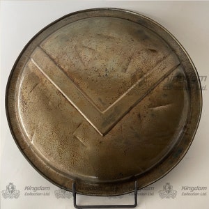 May include: A round, metal shield with a weathered, aged finish. The shield has a distinctive V-shaped design in the center, with a raised edge around the perimeter.