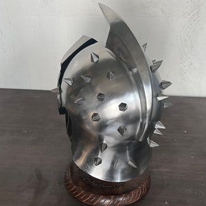 May include: A silver-colored helmet with a prominent central crest and numerous conical spikes. The helmet is displayed on a wooden stand, showcasing its metallic sheen and intricate design. The overall aesthetic suggests a medieval or fantasy theme.