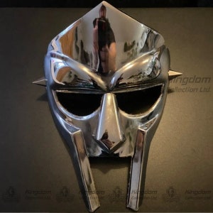 May include: A chrome mask with a distinctive design, featuring a pointed top, angular features, and eye openings. The mask has a reflective surface and is adorned with small, pointed spikes on the sides. The mask is a unique accessory.