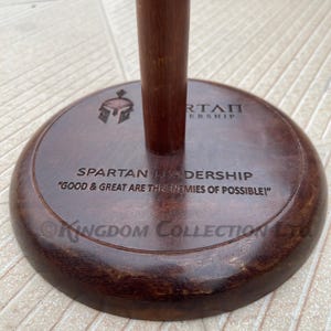 Könnte beinhalten: Ein Holzständer mit dunkelbrauner Oberfläche. Der Ständer hat eine runde Basis mit dem Text "SPARTAN LEADERSHIP" und dem Zitat "GOOD & GREAT ARE THE ENEMIES OF POSSIBLE!" eingraviert. Der Ständer ist dafür ausgelegt, eine Lanze oder ein ähnliches Objekt zu halten.