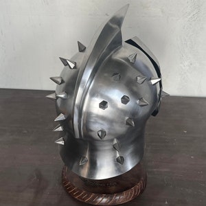 May include: A polished metal helmet with a prominent central ridge and multiple conical spikes. The helmet is mounted on a wooden base, showcasing a medieval or fantasy aesthetic. The metal has a reflective surface.