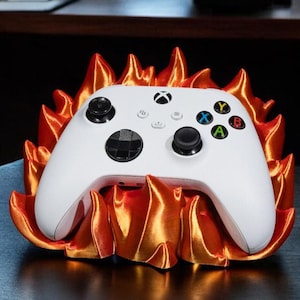 Könnte beinhalten: Ein weißer Xbox-Controller liegt auf einem flammenförmigen Ständer. Der Ständer hat einen Farbverlauf von Orange und Gold. Der Controller hat farbige Knöpfe und schwarze Joysticks. Der Ständer steht auf einer dunklen Oberfläche.