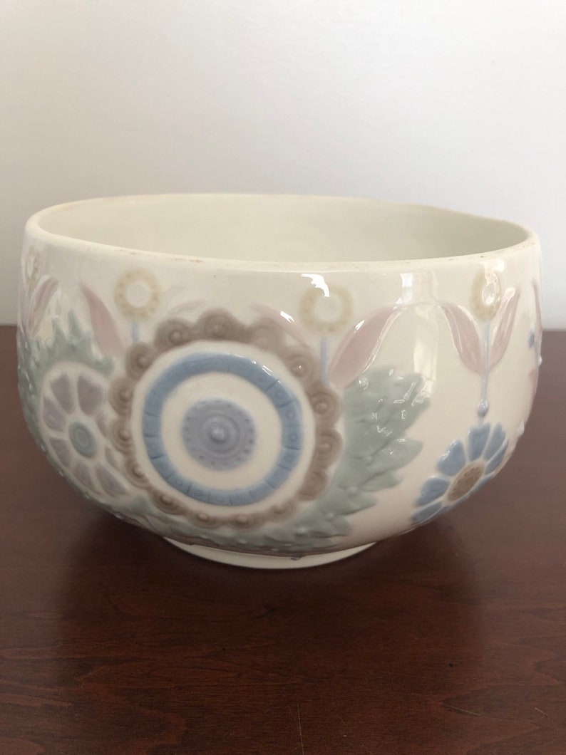 May include: A white ceramic bowl with a raised floral design in shades of blue, green, and brown. The bowl has a rounded shape and a smooth, glossy finish.