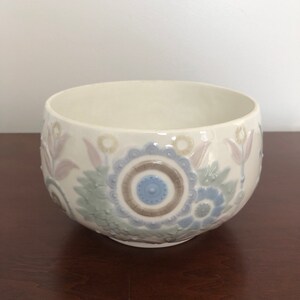 May include: A white ceramic bowl with a floral design in shades of blue, green, and brown. The bowl has a raised design and a glossy finish.