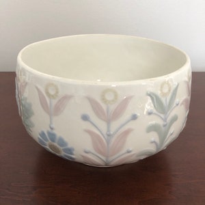 May include: A white ceramic bowl with a floral design in shades of pink, blue, and green. The bowl has a rounded shape and a slightly raised rim.