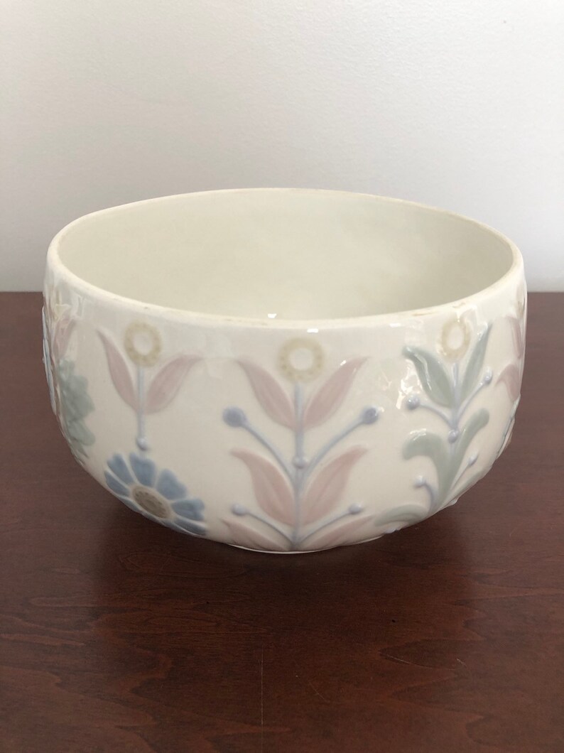 May include: A white ceramic bowl with a floral design in shades of pink, blue, and green. The bowl has a rounded shape and a slightly raised rim.