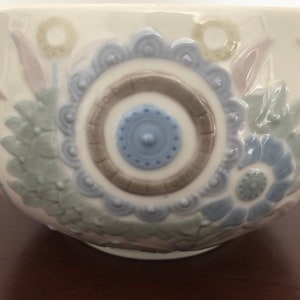 May include: A white ceramic bowl with a floral design. The bowl features a blue and brown circular pattern in the center, surrounded by stylized flowers in shades of blue, green, and pink.