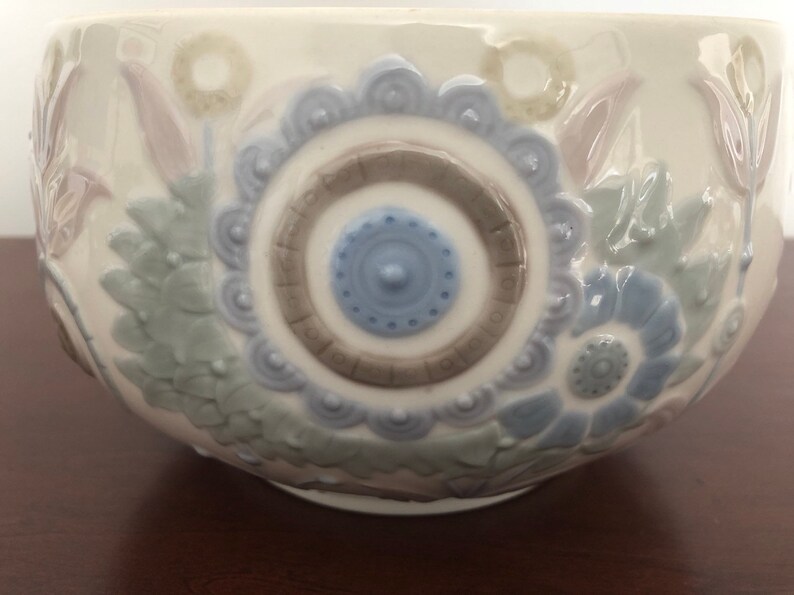 May include: A white ceramic bowl with a floral design. The bowl features a blue and brown circular pattern in the center, surrounded by stylized flowers in shades of blue, green, and pink.