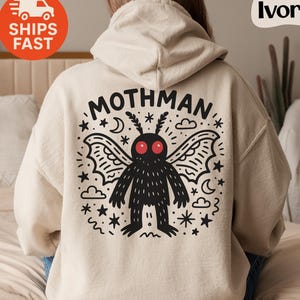 May include: Ivory-colored hoodie with a graphic of the Mothman, a cryptid, on the back. The design features the word "MOTHMAN" above a black creature with red eyes, wings, and stars. The hoodie has a hood and long sleeves.