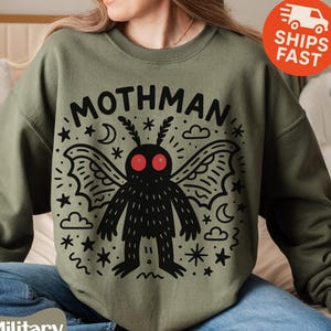 May include: Military green sweatshirt featuring a black Mothman graphic with red eyes, wings, and the word "MOTHMAN" above it. The design includes stars, clouds, and crescent moons. The sweatshirt is a crew neck style.
