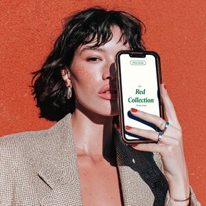 Könnte beinhalten: Ein Smartphone mit goldenem Rahmen zeigt den Text "Red Collection" auf weißem Hintergrund. Die Person hält das Telefon vor einer rot-orangen Wand. Sie trägt einen gemusterten Blazer und silberne Ringe. Der Text enthält auch "Monday Design".