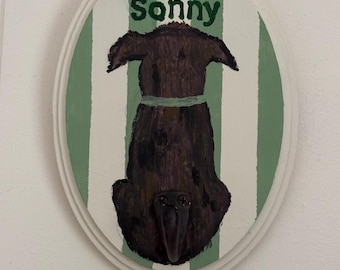 May include: An oval wooden wall plaque featuring a painted dark brown dog seen from behind, wearing a light green collar. The background has vertical sage green and white stripes. The name Sonny is painted in dark green above the dog.