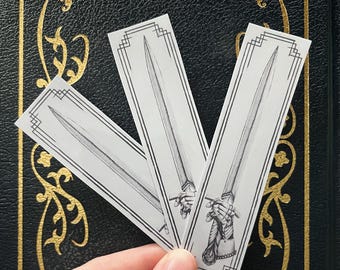 May include: Three white rectangular bookmarks with a black and white illustration of a sword. The sword is held in a hand with detailed line art. The bookmarks have a decorative border.