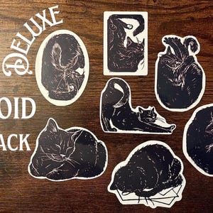 Puede incluir: Una colecci&oacute;n de siete pegatinas de gatos en blanco y negro con varias posturas felinas, incluyendo posiciones durmiendo, estir&aacute;ndose y acurrucadas. El texto DELUXE VOID PACK est&aacute; impreso en blanco en el lado izquierdo de la disposici&oacute;n de pegatinas.