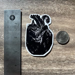 Puede incluir: Una pegatina en blanco y negro con una ilustraci&oacute;n estilizada de un gato enroscado. La pegatina est&aacute; colocada junto a una regla de metal que muestra una altura de unos 6,4 cm y una moneda para comparar el tama&ntilde;o.