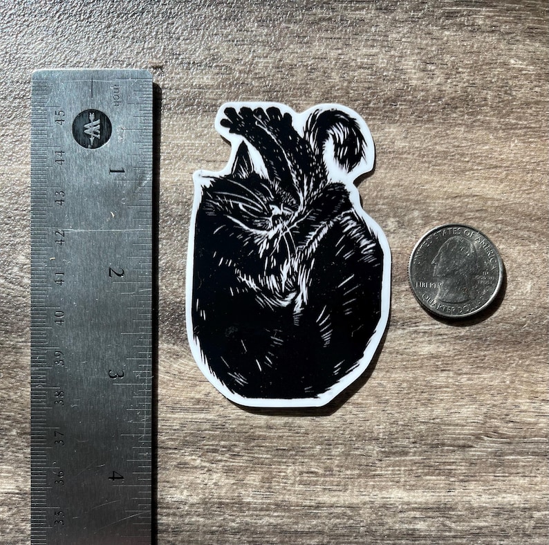 Puede incluir: Una pegatina en blanco y negro con una ilustraci&oacute;n estilizada de un gato enroscado. La pegatina est&aacute; colocada junto a una regla de metal que muestra una altura de unos 6,4 cm y una moneda para comparar el tama&ntilde;o.