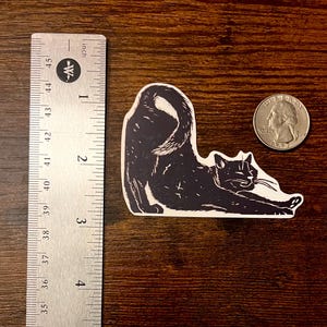 Puede incluir: Una pegatina en blanco y negro de un gato estirando la espalda, colocada junto a una regla de metal que muestra una longitud de unos 8,9 cm y una moneda de cuarto de d&oacute;lar estadounidense para mostrar la escala del tama&ntilde;o de la pegatina.