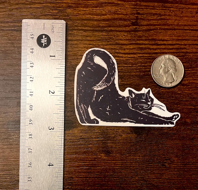 Puede incluir: Una pegatina en blanco y negro de un gato estirando la espalda, colocada junto a una regla de metal que muestra una longitud de unos 8,9 cm y una moneda de cuarto de d&oacute;lar estadounidense para mostrar la escala del tama&ntilde;o de la pegatina.