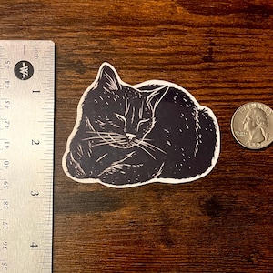 Puede incluir: Una pegatina en blanco y negro de un gato durmiendo con estilo de linograbado, colocada junto a una regla met&aacute;lica que muestra un ancho de aproximadamente 7,6 cm y una moneda de cuarto de d&oacute;lar estadounidense para escala sobre madera.