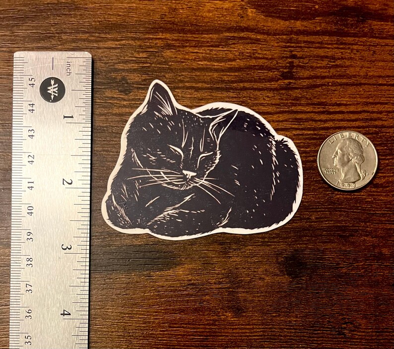 Puede incluir: Una pegatina en blanco y negro de un gato durmiendo con estilo de linograbado, colocada junto a una regla met&aacute;lica que muestra un ancho de aproximadamente 7,6 cm y una moneda de cuarto de d&oacute;lar estadounidense para escala sobre madera.
