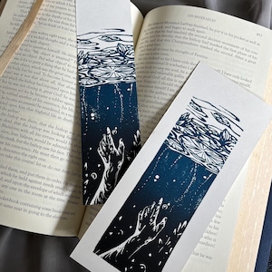 May include: Two black and white linocut prints of a hand reaching up from a dark blue body of water with lily pads on the surface. The prints are on a white background.