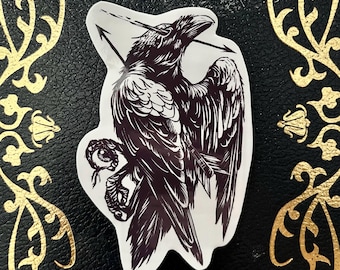 May include: A sticker featuring a detailed illustration of a raven pierced by an arrow. The raven is depicted in shades of brown and black, with intricate feather details. The sticker has a white border and is set against a black background with gold decorative elements.