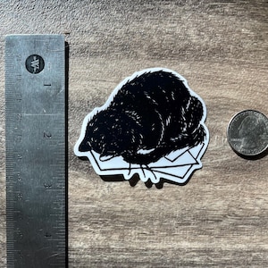 Puede incluir: Una pegatina en blanco y negro con una ilustraci&oacute;n estilizada de un animal oscuro y peludo acurrucado sobre una pila de sobres blancos. Una regla met&aacute;lica en cent&iacute;metros y una moneda est&aacute;n colocadas al lado para mostrar la escala.