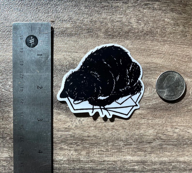 Puede incluir: Una pegatina en blanco y negro con una ilustraci&oacute;n estilizada de un animal oscuro y peludo acurrucado sobre una pila de sobres blancos. Una regla met&aacute;lica en cent&iacute;metros y una moneda est&aacute;n colocadas al lado para mostrar la escala.