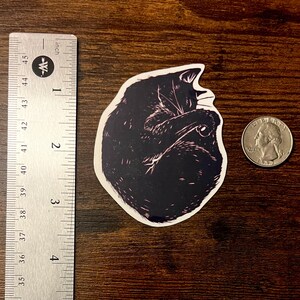 Puede incluir: Una pegatina troquelada que muestra un gato negro enroscado en forma de bola con detalles de l&iacute;neas blancas. La pegatina est&aacute; colocada junto a una regla met&aacute;lica que muestra una altura de unos 6,4 cm y una moneda de 25 centavos como referencia.