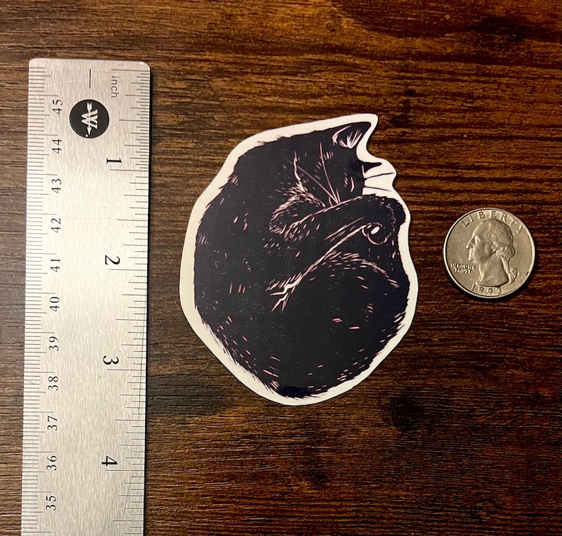 Puede incluir: Una pegatina troquelada que muestra un gato negro enroscado en forma de bola con detalles de l&iacute;neas blancas. La pegatina est&aacute; colocada junto a una regla met&aacute;lica que muestra una altura de unos 6,4 cm y una moneda de 25 centavos como referencia.