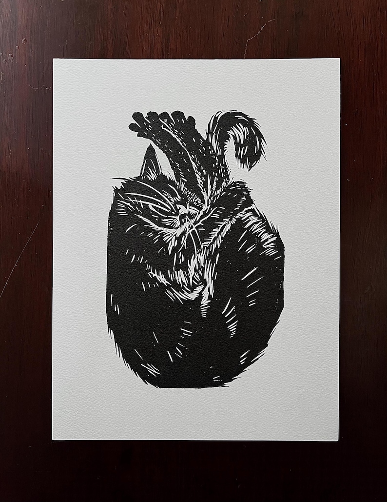 May include: Black and white linocut print of a cat curled up sleeping. The cat is depicted in a simple, stylized manner, with its fur rendered in a series of parallel lines.
