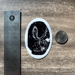 Puede incluir: Una pegatina ovalada con una ilustraci&oacute;n en blanco y negro de un gato enroscado. Una regla de metal muestra que la pegatina mide aproximadamente 6,4 cm de alto, y una moneda est&aacute; colocada al lado para una clara comparaci&oacute;n de tama&ntilde;o.