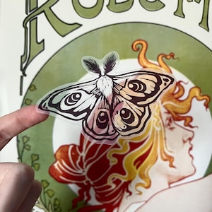 May include: A transparent sticker of a moth with detailed wing patterns in shades of brown, pink, and black. The moth is held in front of a vintage-style poster with green and red accents and the name "ROBERT" in large letters.