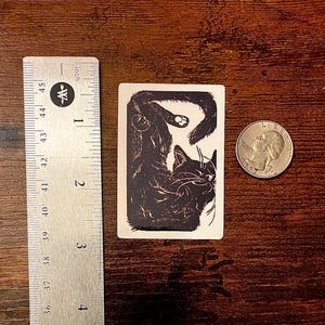 Puede incluir: Una pegatina rectangular con una ilustraci&oacute;n en blanco y negro de un gato enroscado. La pegatina est&aacute; situada junto a una regla met&aacute;lica que muestra una altura de unos 6,4 cm y una moneda para referencia de escala.