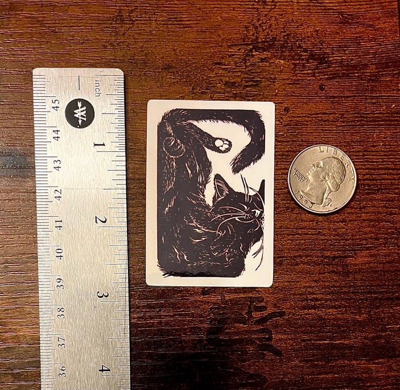 Puede incluir: Una pegatina rectangular con una ilustraci&oacute;n en blanco y negro de un gato enroscado. La pegatina est&aacute; situada junto a una regla met&aacute;lica que muestra una altura de unos 6,4 cm y una moneda para referencia de escala.