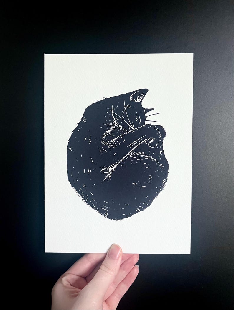 Op de afbeelding: Zwart-wit linoleumsnede van een opgerolde kat. De kat is afgebeeld in een gestileerde, grafische stijl, met gedetailleerde lijnen die textuur cre&euml;ren. De prent staat op wit papier, vastgehouden door een hand, tegen een zwarte achtergrond.