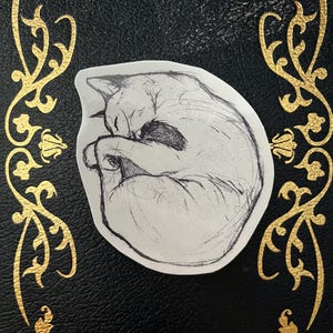 May include: A sticker featuring a curled-up cat sketch in black and white. The cat is drawn in a simple, artistic style, with its paws tucked in. The sticker is on a black surface with gold decorative accents.