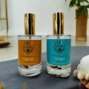 May include: Two glass bottles with gold lids and labels that say "Enchant Mists" and contain crystal infused yoga mat cleaner. The bottles are labeled "Enlighten" and "Ground".