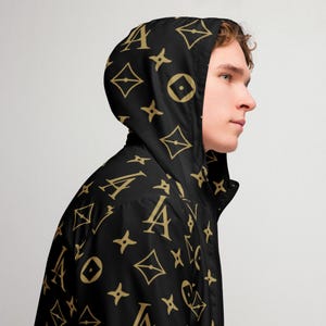 May include: Black hooded jacket with a repeating gold pattern. The pattern includes stylized letters, diamonds, and star-like shapes. The jacket has a zipper closure and a hood.