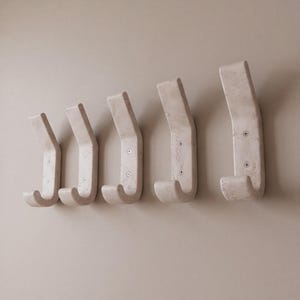 May include: Five off-white, curved wall hooks are mounted on a light beige wall. Each hook has a rectangular top and a curved hook shape. The hooks are made of a textured material and are evenly spaced.