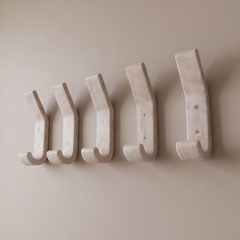 May include: Five off-white, curved wall hooks are mounted on a light beige wall. Each hook has a rectangular top and a curved hook shape. The hooks are made of a textured material and are evenly spaced.
