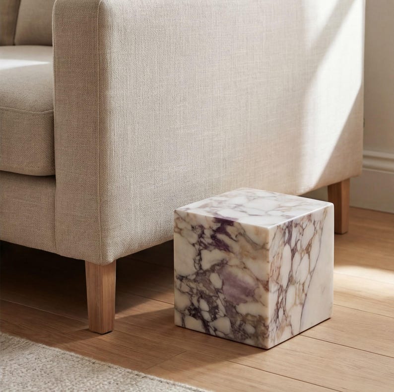 May include: A cube-shaped side table made of marble, featuring a white and purple veined design. It stands on a wooden floor next to a beige sofa. The table's dimensions are approximately 30 cm x 30 cm.