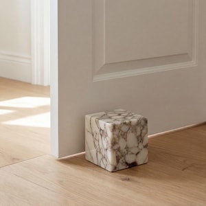 May include: A square marble doorstop with a white and brown patterned design. The doorstop is positioned on a light-coloured wooden floor, near a white door and door frame. The doorstop is a decorative and functional home decor item.