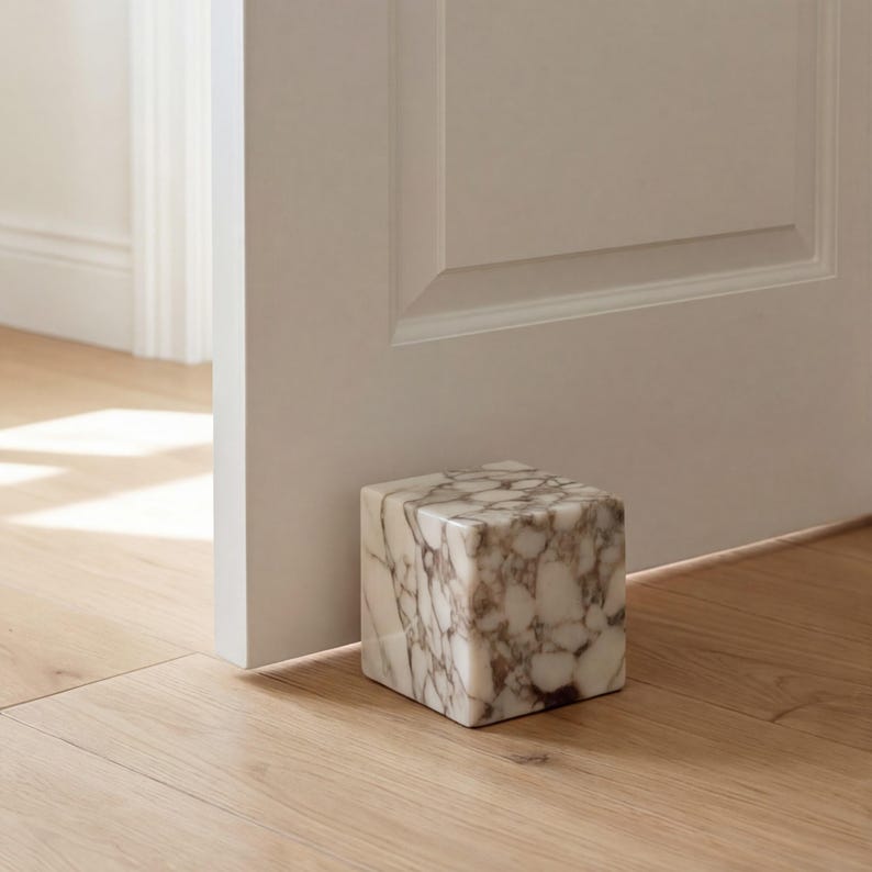 May include: A square marble doorstop with a white and brown patterned design. The doorstop is positioned on a light-coloured wooden floor, near a white door and door frame. The doorstop is a decorative and functional home decor item.