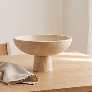 Puede incluir: Un cuenco beige sobre pedestal con una base ancha y poco profunda. El cuenco est&aacute; hecho de un material texturizado y se asienta sobre una base cil&iacute;ndrica corta. Una servilleta de lino beige doblada descansa sobre la mesa de madera en primer plano.