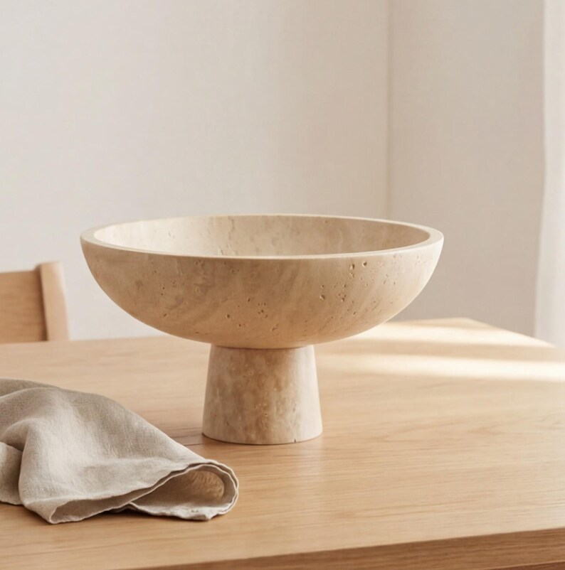 Puede incluir: Un cuenco beige sobre pedestal con una base ancha y poco profunda. El cuenco est&aacute; hecho de un material texturizado y se asienta sobre una base cil&iacute;ndrica corta. Una servilleta de lino beige doblada descansa sobre la mesa de madera en primer plano.