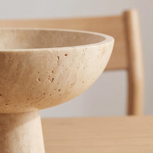 Puede incluir: Un cuenco beige texturizado sobre pedestal con una base ancha y poco profunda. El cuenco est&aacute; hecho de un material similar a la piedra y tiene forma redondeada. El fondo incluye una silla y una mesa de madera.